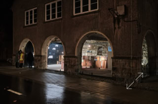 Ein Gebäude mit drei offenen Bögen, durch die ein beleuchteter Innenraum mit einer Installation aus Kartons sichtbar ist; draußen stehen drei Personen bei Nacht.