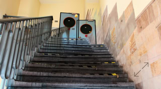 Zwei versetzt übereinander geblendete Bilder der gleichen Treppe führen oben zu einer Tür mit Bullauge, Geländer links, Wand mit Fliesen rechts. Tennisbälle springen die Treppe herunter.