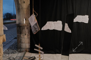 Kunstinstallation mit hängenden Papieren und einem Spiegel vor einem schwarzen Hintergrund, daneben ein offener Torbogen mit Blick nach draußen.