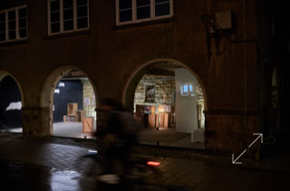 Ein verschwommener Radfahrer fährt nachts vor einem Gebäude mit beleuchteten Bögen und Schaufenstern vorbei.